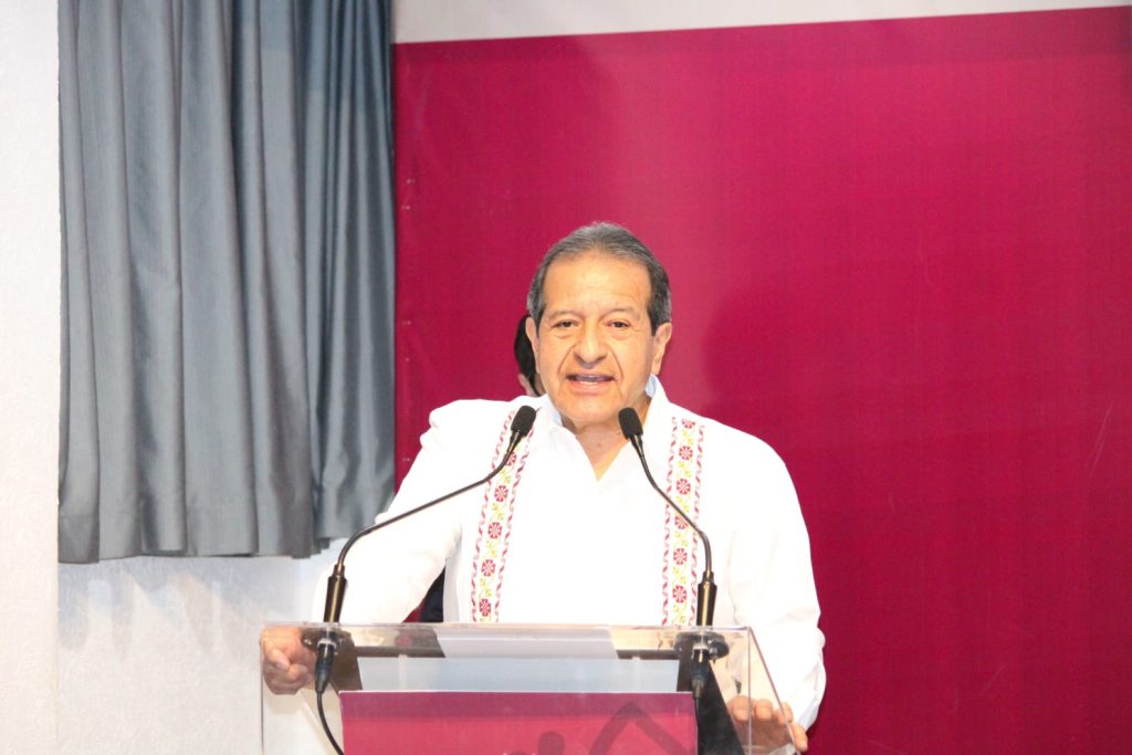 Hombre hablando en un podio durante una conferencia o evento formal, vestido con camisa blanca y detalles bordados, frente a un fondo de cortinas grises y pared rosa.