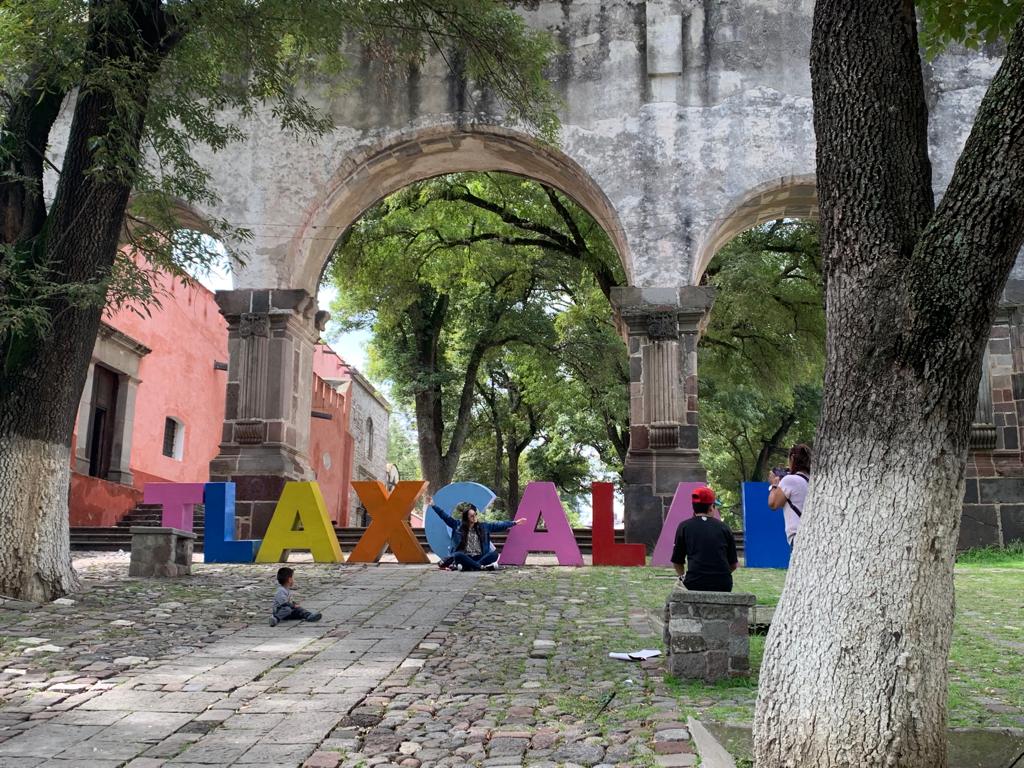 Entrada histórica en Tlaxcala con arco de piedra y letras grandes que forman "TLAXCALA" en un parque con árboles y personas disfrutando del lugar.