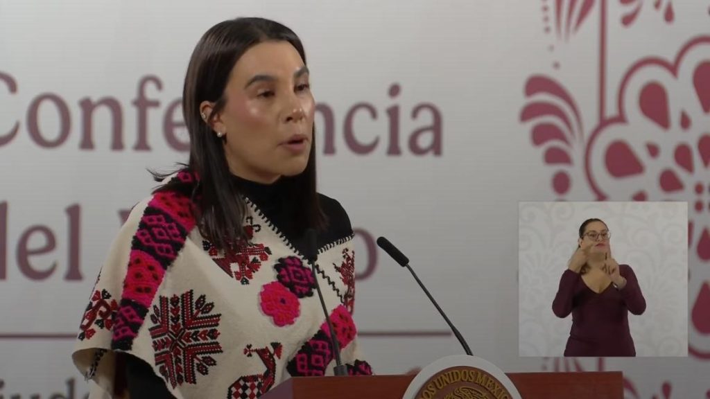 La mujer en la conferencia de prensa habla en un micrófono, vistiendo un suéter con patrones tradicionales en colores rosa, negro y blanco, en un evento formal en Tlaxcala, México.
