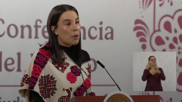 La mujer en la conferencia de prensa habla en un micrófono, vistiendo un suéter con patrones tradicionales en colores rosa, negro y blanco, en un evento formal en Tlaxcala, México.
