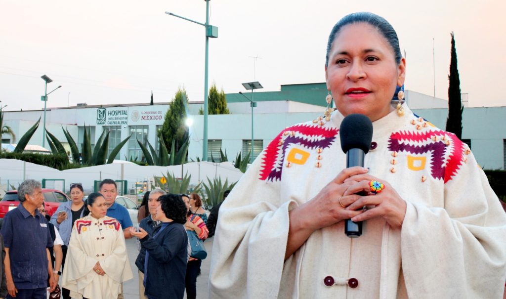 Una mujer indígena hablando en un micrófono durante un evento público en Tlaxcala, México, con varias personas y un hospital en el fondo.