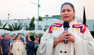 Una mujer indígena hablando en un micrófono durante un evento público en Tlaxcala, México, con varias personas y un hospital en el fondo.