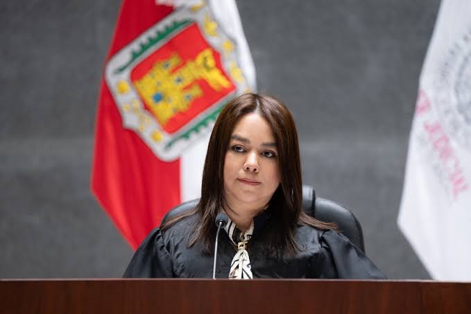 Juez en una sala de tribunal con bandera de Tlaxcala y bandera institucional en el fondo, vestida con toga negra y blusa blanca, en una audiencia judicial.