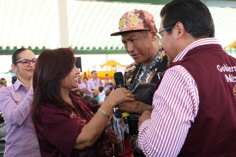 Cuidado de la salud en Tlaxcala: una mujer entrega un reconocimiento a un funcionario durante un evento oficial en un espacio abierto con estructura metálica y varias personas observando.
