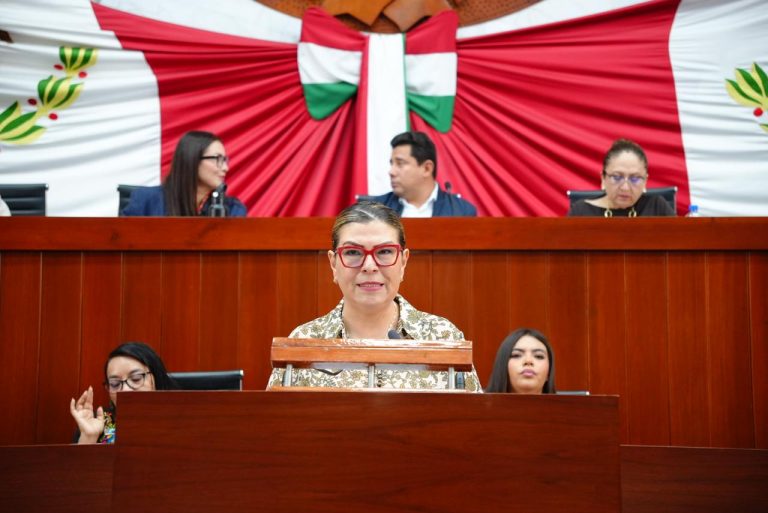 Una mujer en un podio en una sesión del ayuntamiento, con fondo decorado con banderas de México y Tlaxcala, y varias personas sentadas en la mesa del consejo.