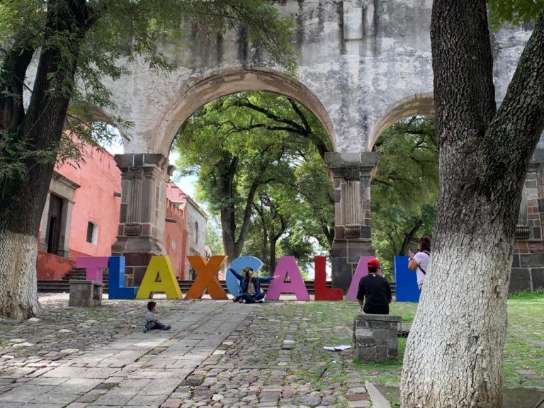 Árboles y estructura histórica en el centro de Tlaxcala, con letras grandes que forman la palabra "Tlaxcala" en colores vibrantes, en un día nublado.