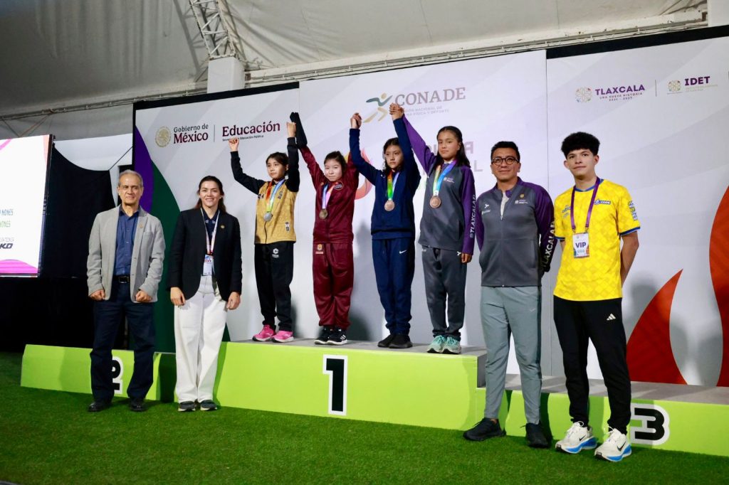Niños en el podio de premiación en una competencia deportiva en Tlaxcala, con medallas y trofeos, celebrando sus logros en un evento oficial.