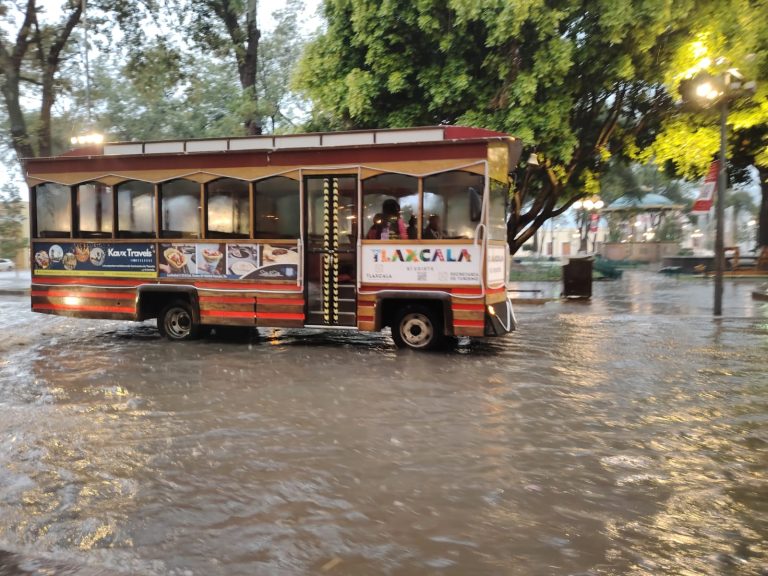 Tranvía turístico de Tlaxcala en una calle inundada por la lluvia, con árboles y luces en el fondo, durante un día lluvioso en la ciudad.