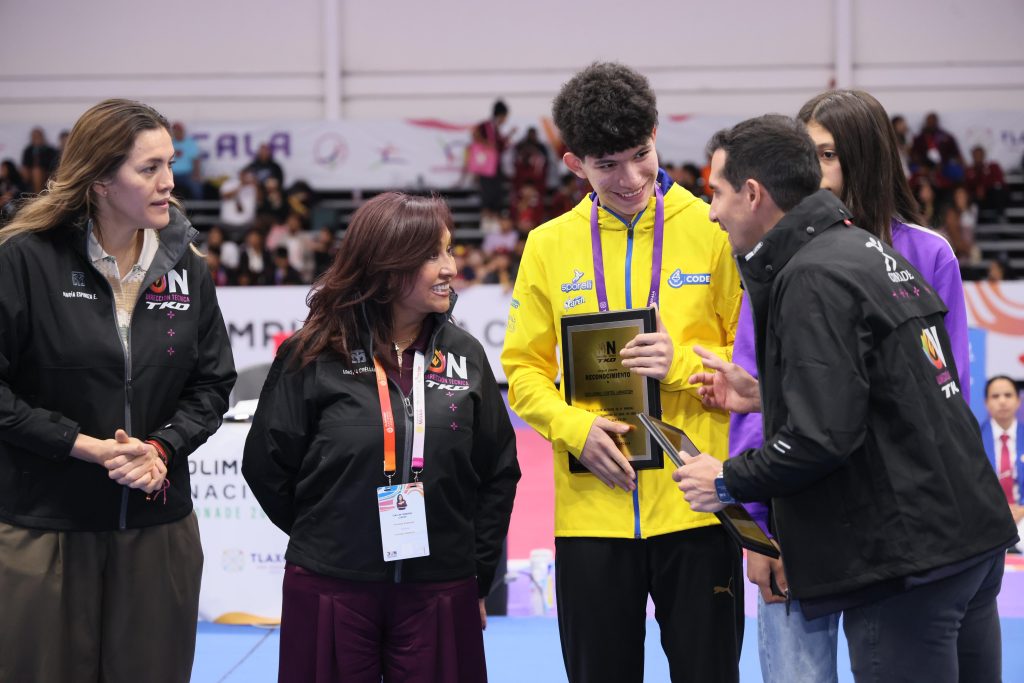 Reconocimiento de medallas en un evento deportivo, con jóvenes atletas y oficiales en un entorno de competencia y premiación en un gimnasio con espectadores en el fondo.