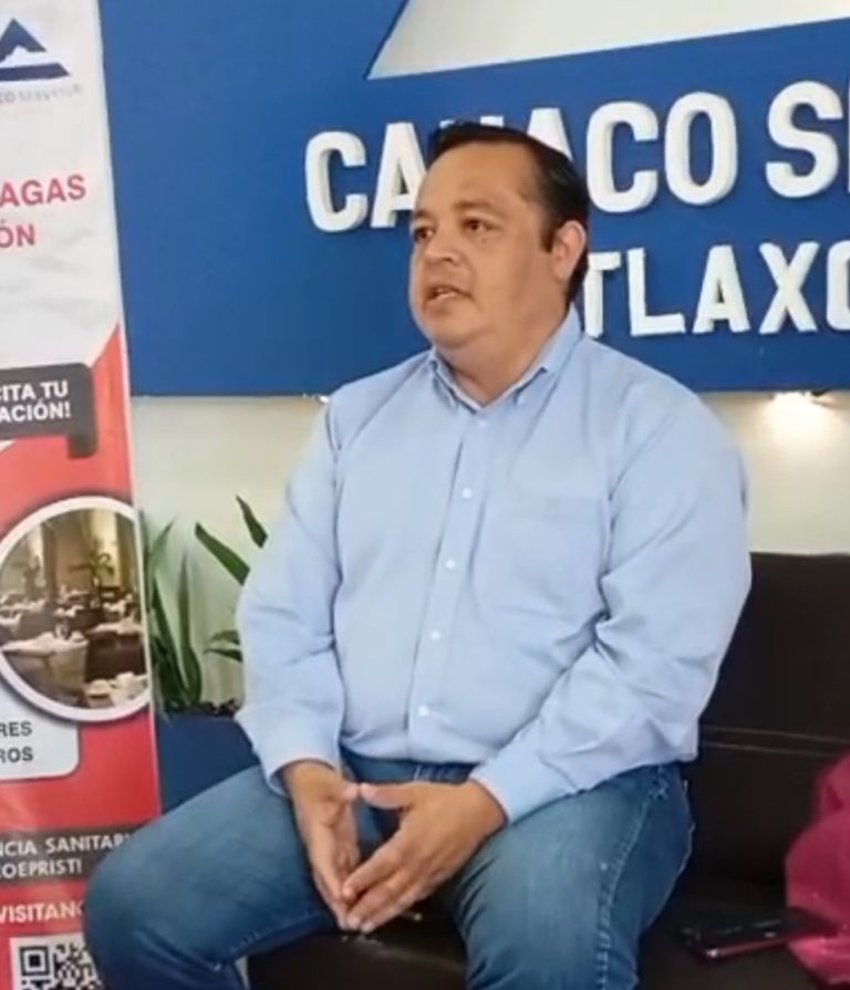 Hombre sentado en una entrevista o conferencia en un espacio con señalización y plantas decorativas.