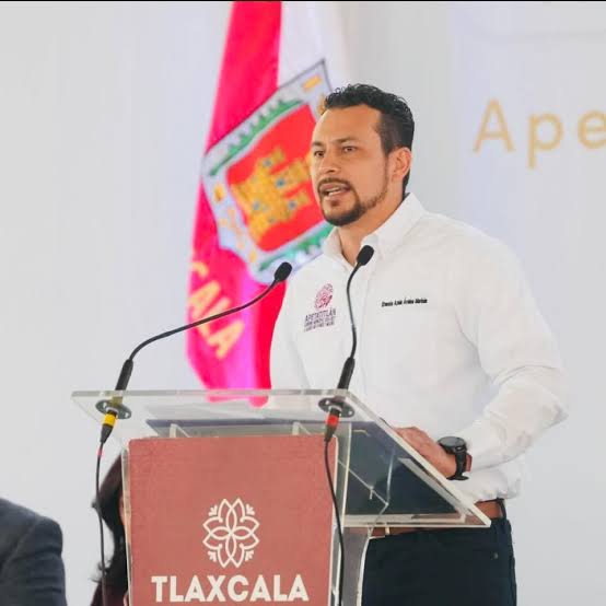 El Periódico de Tlaxcala: Hombre hablando en un podio con bandera de Tlaxcala en el fondo, vestido con camisa blanca y barba, en un evento oficial.