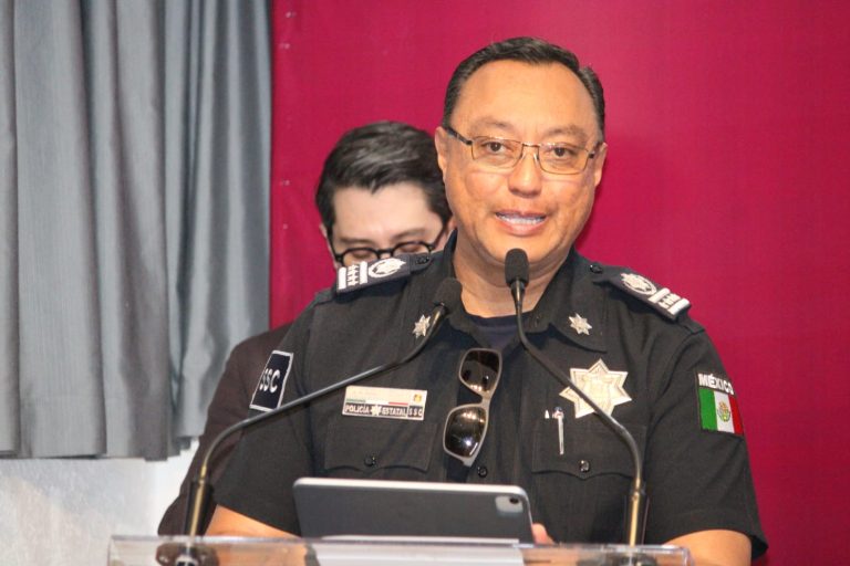 Oficial de policía en uniforme durante una conferencia de prensa en un evento oficial, con fondo de cortinas grises y rojas.