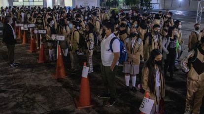 Estudiantes en fila en un evento escolar, usando mascarillas y manteniendo distancia social, en un espacio al aire libre con iluminación artificial y señalización visible.