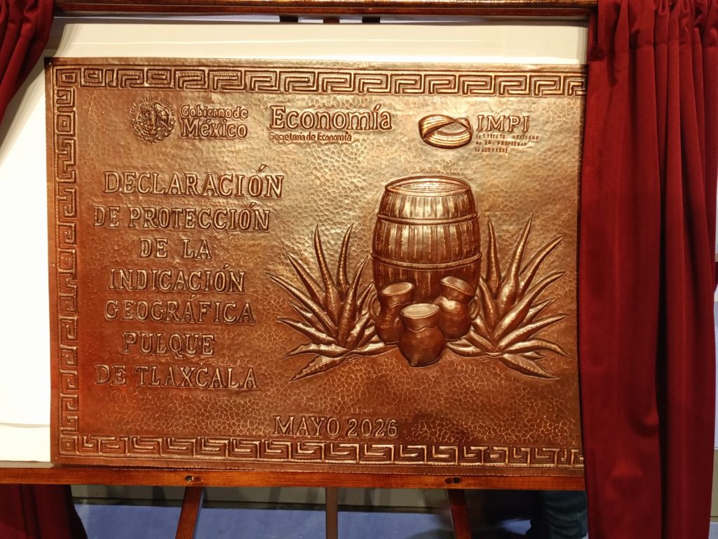 Declaración de protección de la indicación geográfica pulque de Tlaxcala, mayo 2026, en un panel de cobre con símbolos tradicionales y un fondo de cortinas rojas.