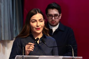 Mujer hablando en un podio con micrófono, acompañada por un hombre en el fondo, en un escenario con fondo rojo.