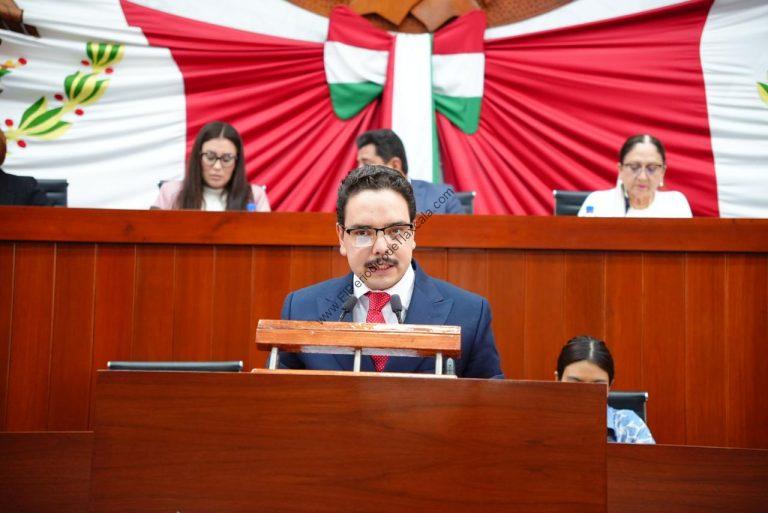 Hombre en un podio en un evento oficial con fondo decorado en colores de la bandera mexicana.