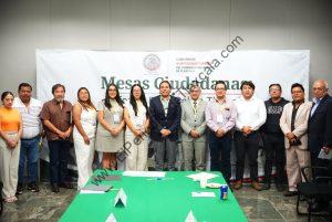 Grupo de personas en una reunión oficial en un espacio con fondo de cartel que dice "Mesas Ciudadanas" y logotipo de la Comisión de Asuntos Electorales del Congreso del Estado de Tlaxcala.
