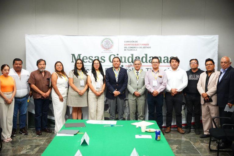 Grupo de personas en una reunión oficial en un espacio con fondo de cartel que dice "Mesas Ciudadanas" y logotipo de la Comisión de Asuntos Electorales del Congreso del Estado de Tlaxcala.