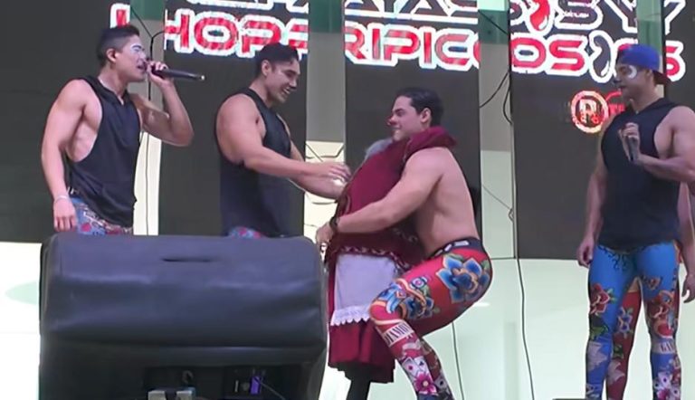 Hombre en un escenario con otros tres hombres, uno de ellos con un micrófono, participando en un evento de lucha libre o espectáculo, con fondo de pantallas y luces.