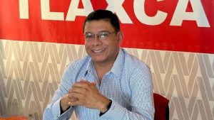 El hombre sonriente con gafas y camisa a cuadros en un entorno con cartel rojo y fondo decorativo.