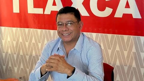 El hombre sonriente con gafas y camisa a cuadros en un entorno con cartel rojo y fondo decorativo.