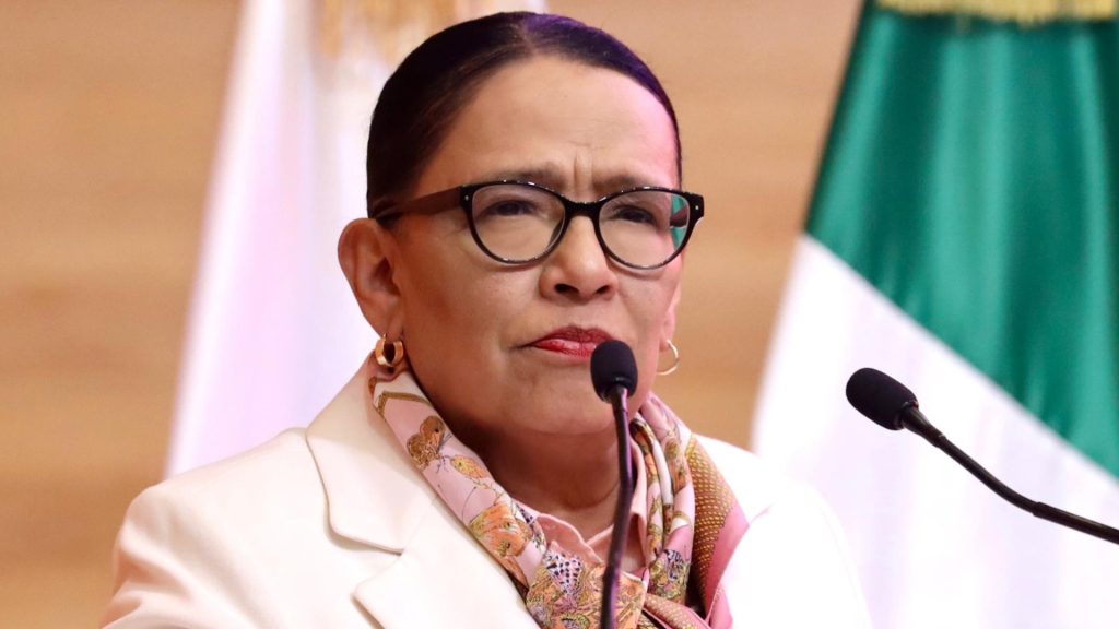 Una mujer con gafas y peinado recogido hablando en un micrófono durante una conferencia o evento oficial, con banderas mexicanas de fondo.