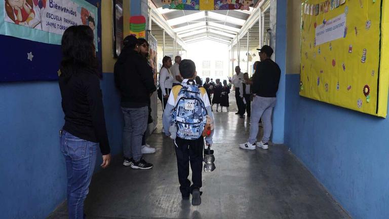 Estudiante con mochila en pasillo escolar, rodeado de otros alumnos y profesores en un ambiente educativo en Tlaxcala, México.